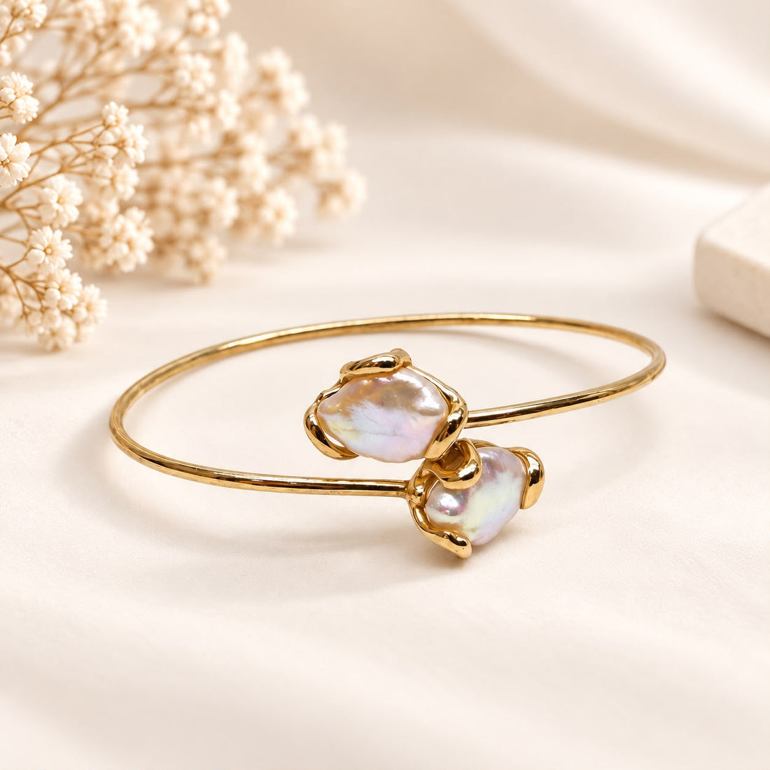 Bracciale rigido flessibile dorato in argento 925 con due perle barocche naturali, fotografato frontalmente su sfondo crema.